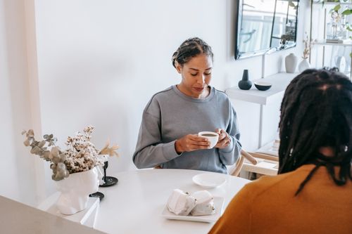 Free Pensive Hispanic female in casual clothes drinking cup of coffee while spending time with friend in light room Stock Photo