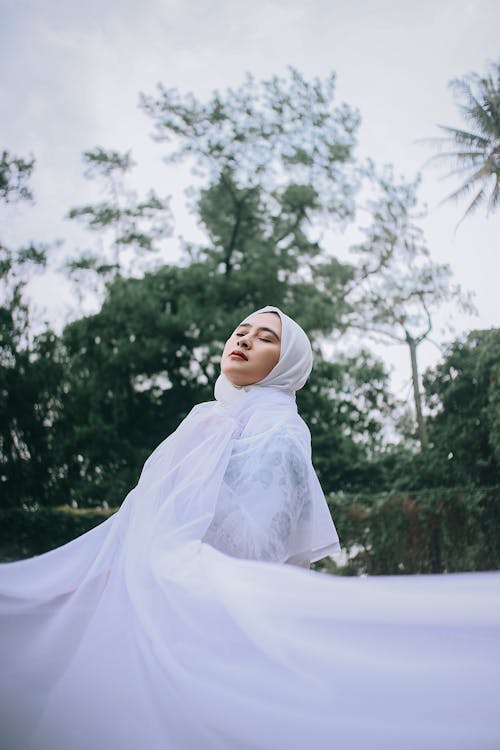 Peaceful female with closed eyes in hijab and traditional wedding dress in green park