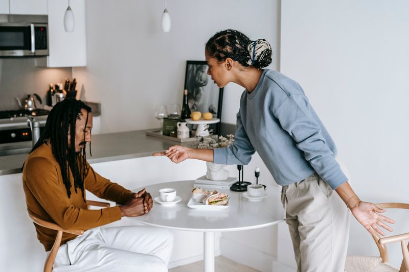 Young black woman blaming ethnic depressed boyfriend sitting at table in living room at home