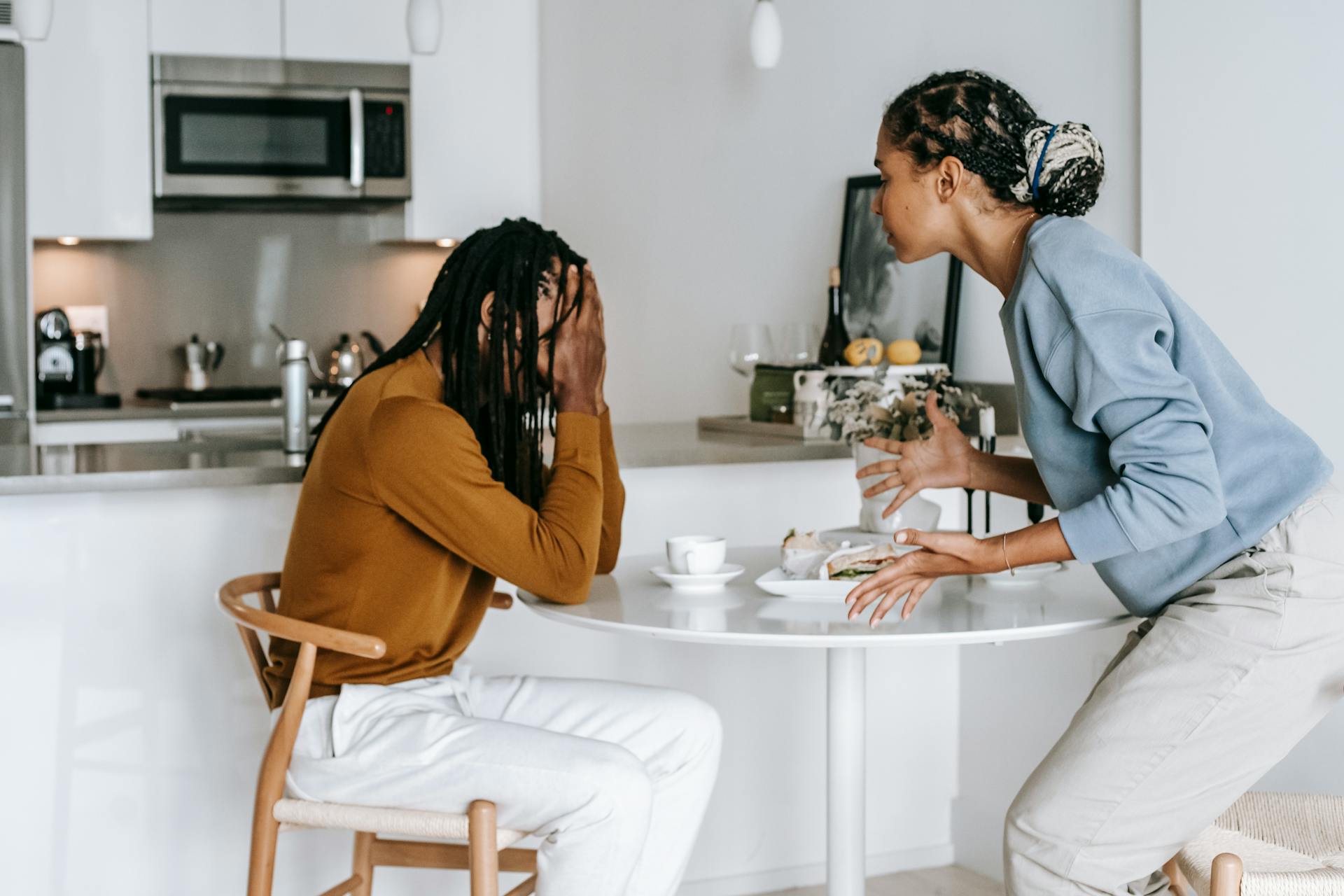 Young black man sitting at table with hands on head and having conflict with standing near table woman in light kitchen