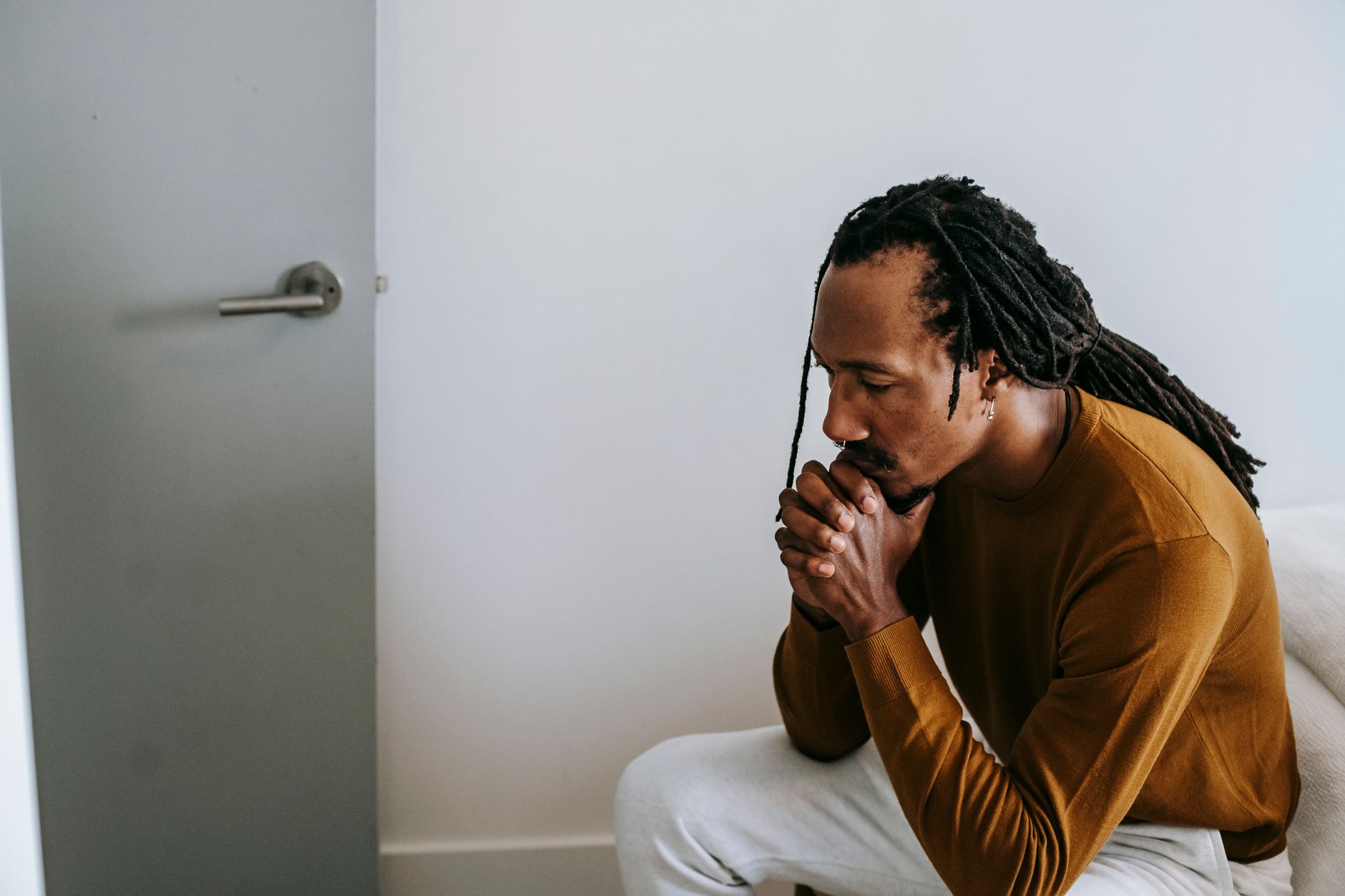 High angle of serious young black man in casual outfit sitting near bed and thinking