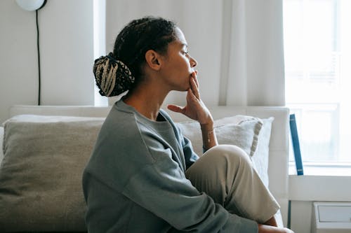 Side view young African American woman sitting on comfortable white bed and thinking in casual clothes in daytime