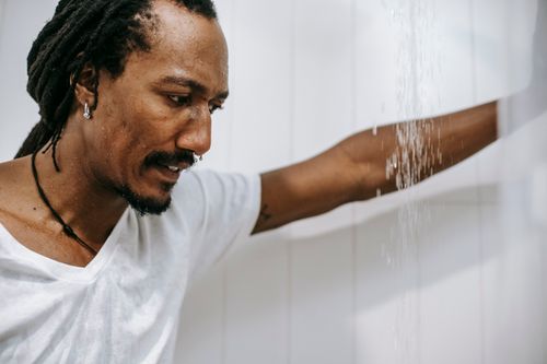 Free Side view of crop young worried African American man with dreadlocks in white t shirt leaning hand on tiled wall while standing under water stream in bathroom Stock Photo