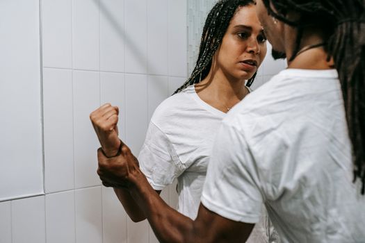 Back view of crop anonymous bearded black man in white t shirt with Afro dreadlocks holding wrist on sad young girlfriend during dispute in bathroom