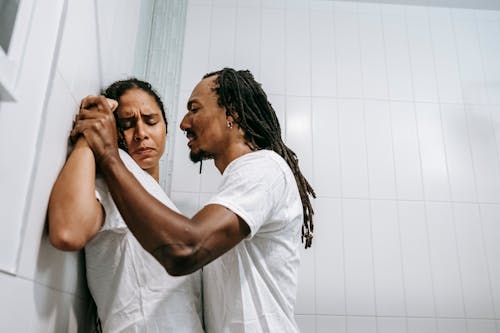 Free Side view of young black angered man with Afro braids in white t shirt pushing sad wife with closed eyes against tiled wall during conflict in bathroom Stock Photo