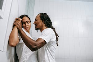 Side view of young black angered man with Afro braids in white t shirt pushing sad wife with closed eyes against tiled wall during conflict in bathroom
