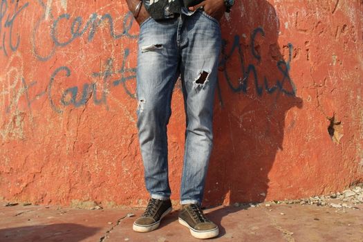 Close-up of a person's legs in ripped jeans and sneakers against a graffitied wall.