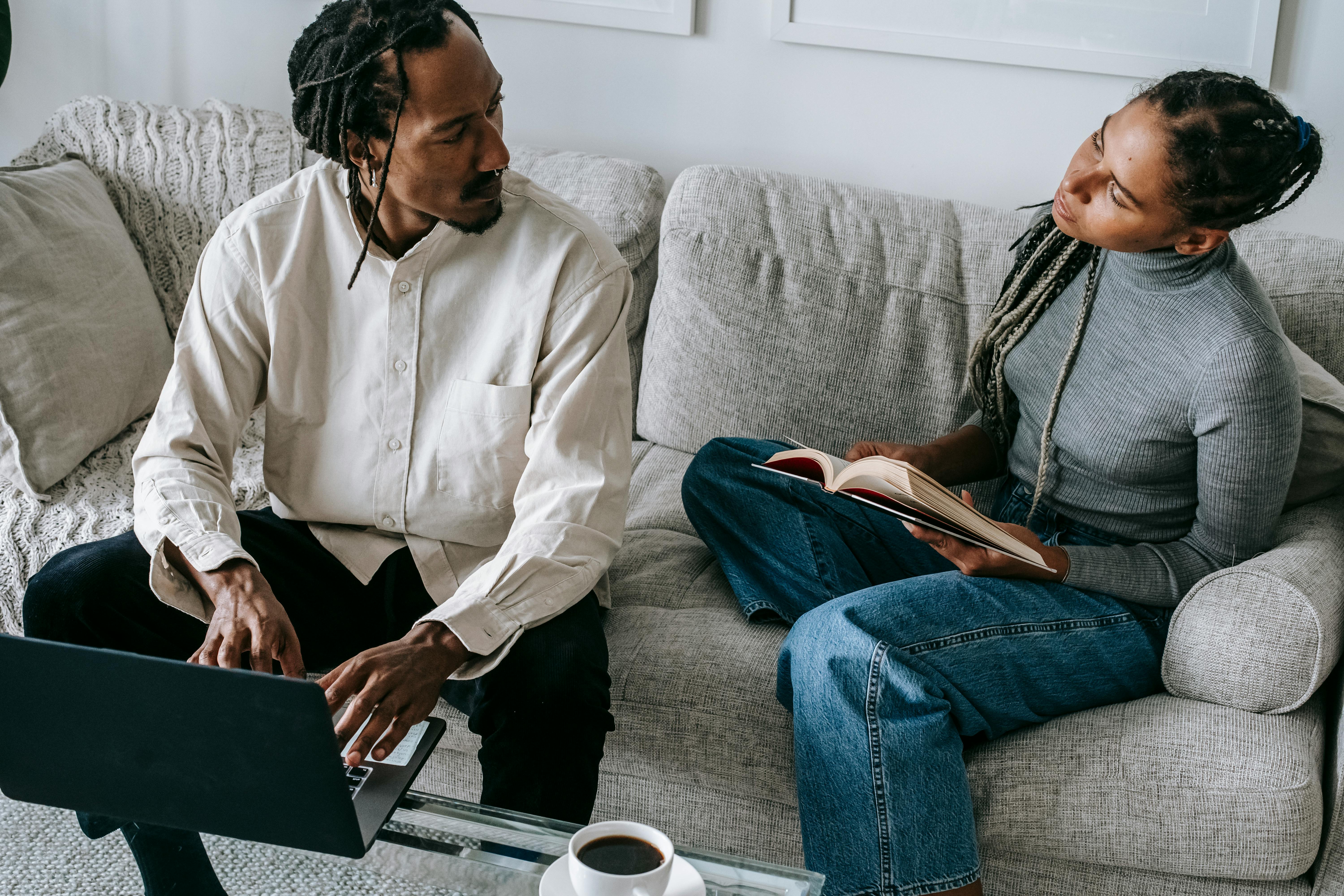 trendy young black couple chatting while sitting on couch with book and laptop