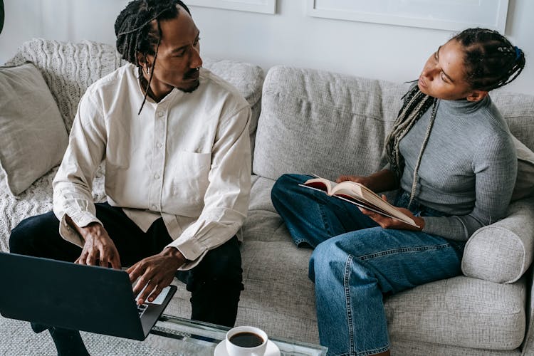 Trendy Young Black Couple Chatting While Sitting On Couch With Book And Laptop