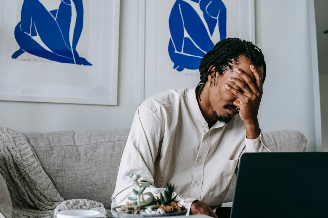 Free Frustrated young bearded African American male freelancer with dreadlocks in casual shirt working on laptop at home and covering eyes with hand after failure Stock Photo