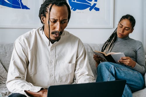 Serious African American man working on laptop sitting on sofa near reading wife
