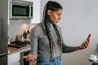 Serious young black lady with Afro braids in casual clothes gesticulating while having unpleasant conversation via video chat on smartphone in modern kitchen