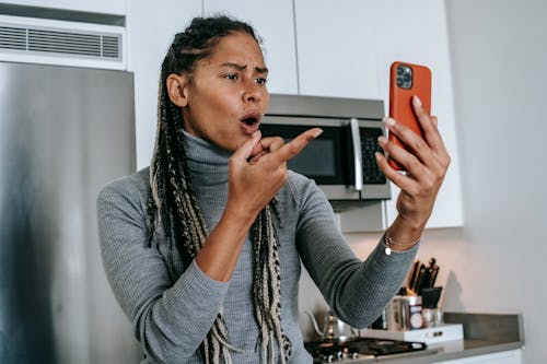 Angry young African American female with long braids in casual clothes pointing at screen nervously while disputing during video call on mobile phone in kitchen