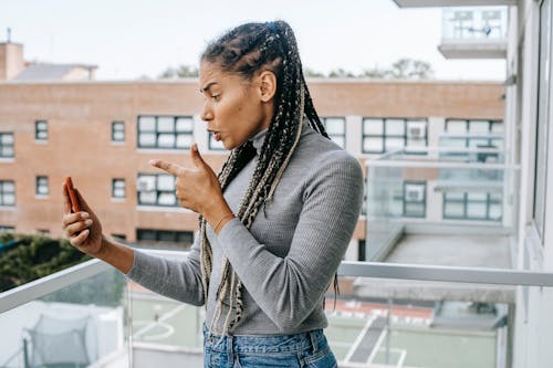 Side view of young furious African American woman with braids in casual outfit pointing at smartphone screen while arguing during video call standing on balcony