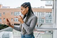 Side view of young furious African American woman with braids in casual outfit pointing at smartphone screen while arguing during video call standing on balcony