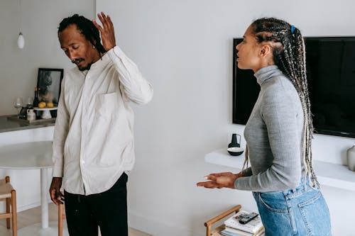 Tired young ethnic man with dreadlocks in casual outfit gesticulating while having argument with wife at home