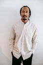一位穿着休闲装、表情严肃的非裔美国男性，手插口袋站立在白色背景前，注视着镜头