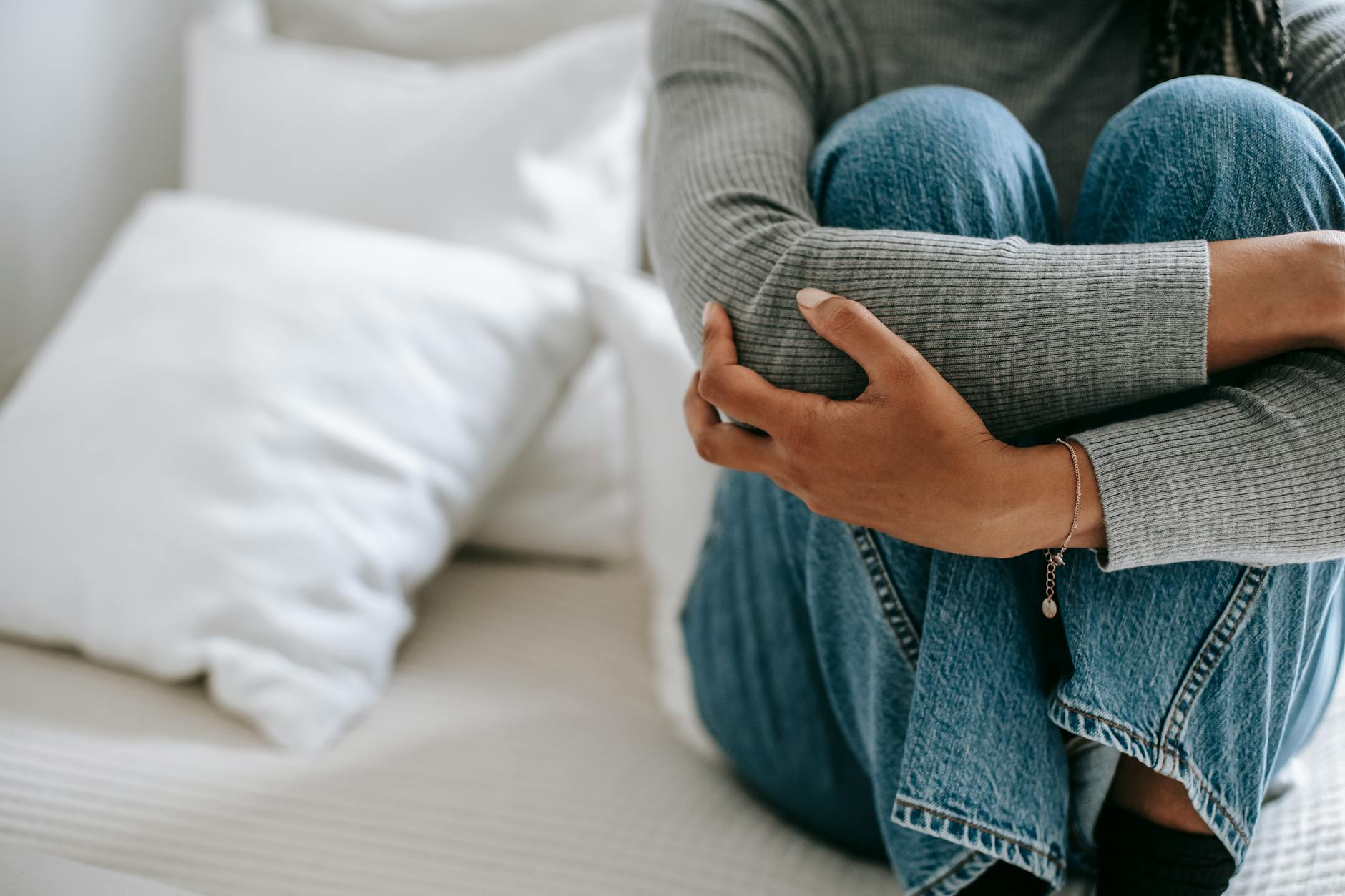 Crop faceless female feeling helpless and frustrated embracing knees while sitting on bed in loneliness