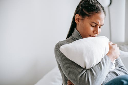 Free Young ethnic female with closed eyes and sad face expression sitting on bed and embracing pillow Stock Photo