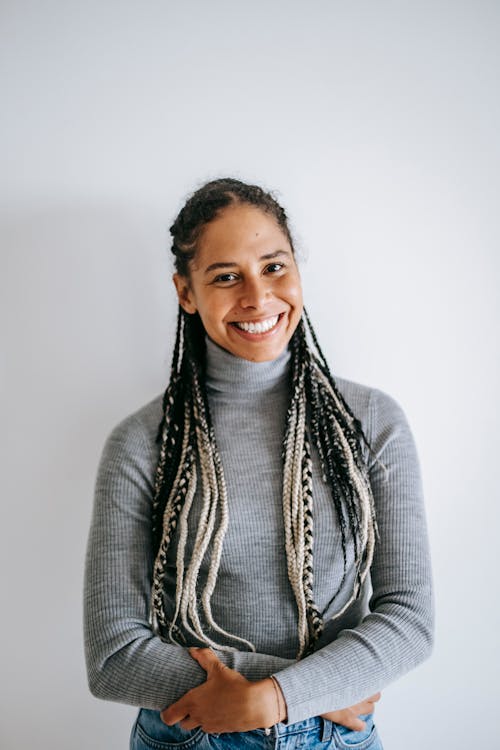 Smiling ethnic female with long braids in turtleneck and jeans crossing hands standing against white background
