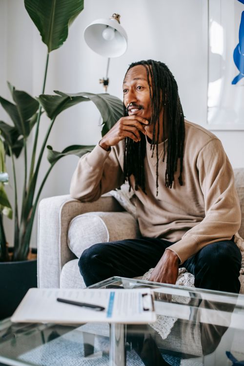 Free Young attentive African American male with dreadlocks looking forward on soft couch in living room Stock Photo