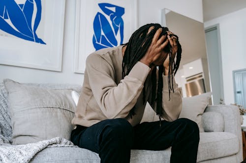 Anonymous desperate African American male with dreadlocks suffering from headache on sofa in living room