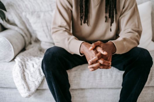 Crop anonymous pensive ethnic male in casual clothes resting on soft couch in living room