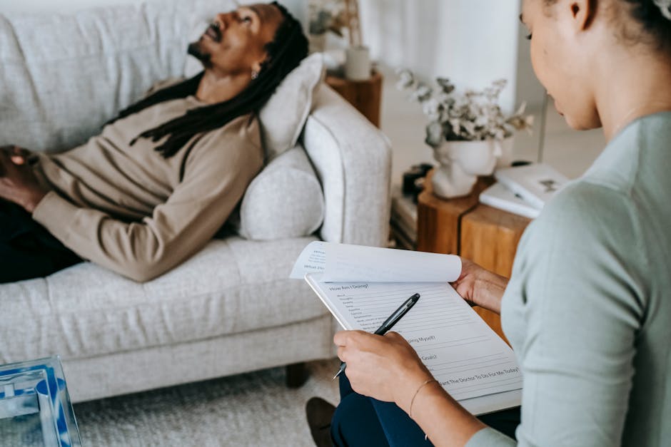 A therapist with clipboard assists a man during a home therapy session on a sofa.