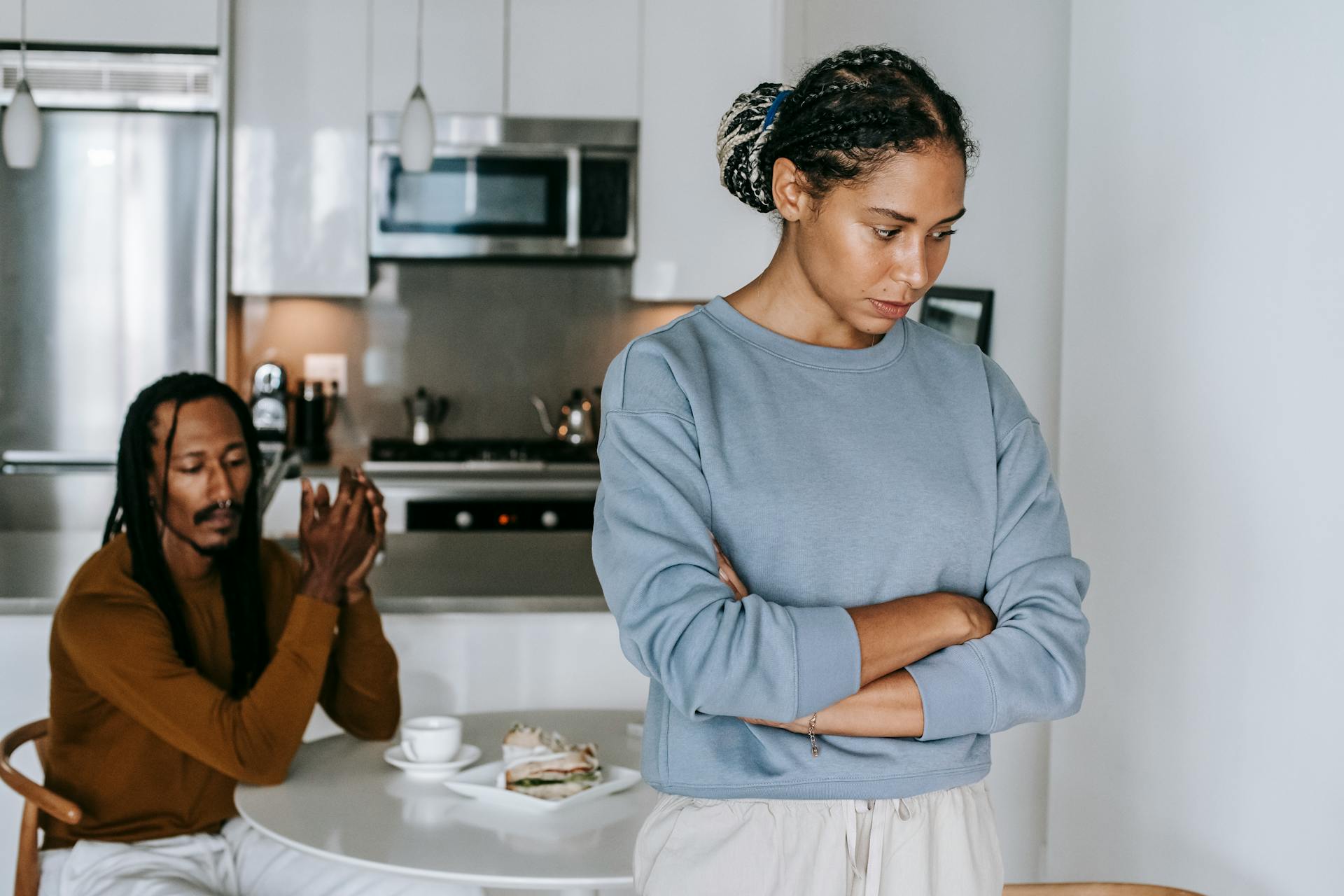 Young discontented African American female with folded arms against male partner at kitchen table during quarrel