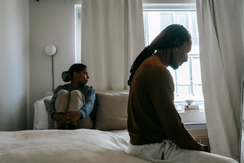 Young frustrated black man with dreadlocks near unhappy girlfriend sitting on bed during conflict