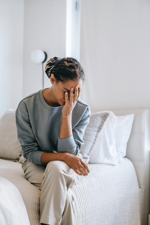 Young unhappy African American female touching face while sitting with crossed legs on bed at home