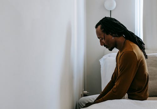 Side view of young sad African American male with dreadlocks sitting on bed and looking down in house