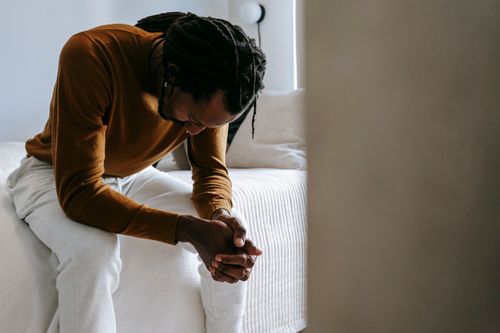 Free Concerned unhappy African American male in casual outfit sitting on bed with hands clasped nervously at home Stock Photo