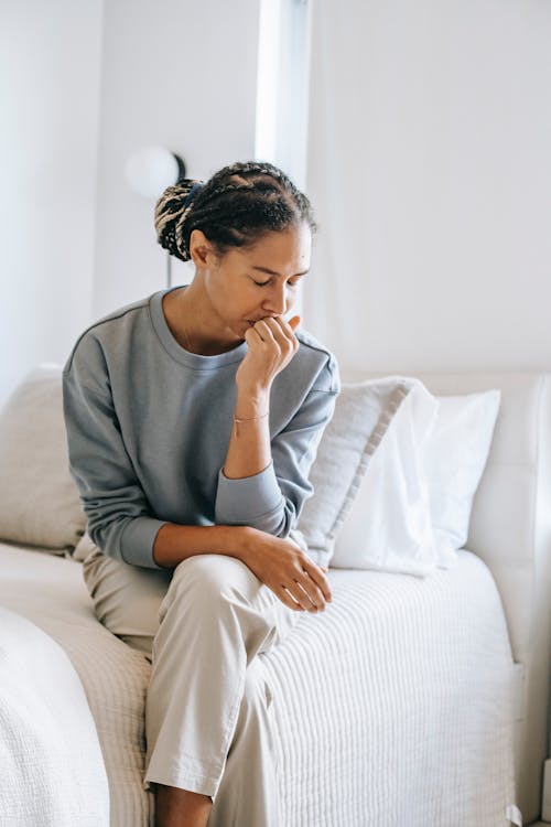 Free Sorrowful crying African American female sitting with eyes closed and crossed legs on bed and touching lips in stress Stock Photo
