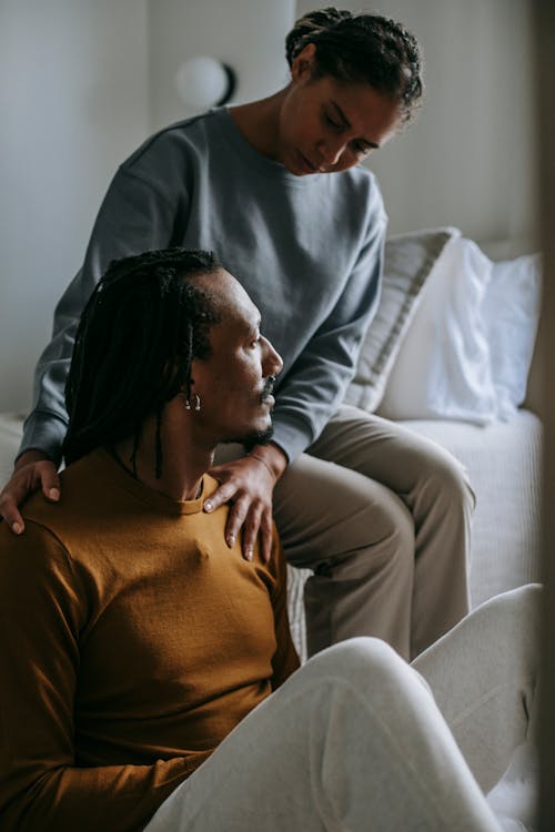 Attentive loving African American female supporting and touching shoulders of stressed frustrated husband while sitting in light bedroom