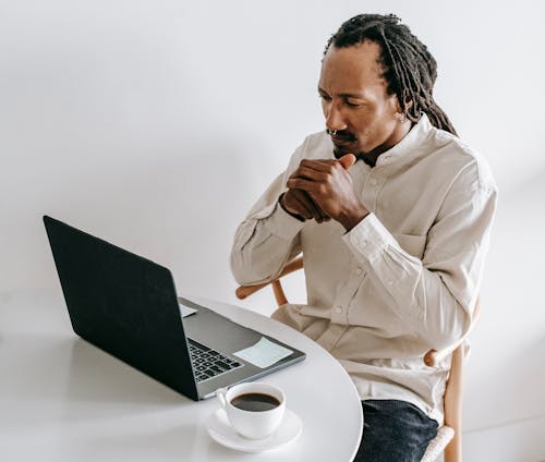 Free Concentrated adult African American male in formal wear sitting at table with cup of coffee and watching video on netbook in light room Stock Photo