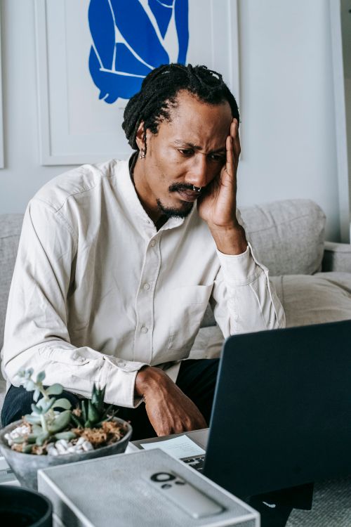 Free Serious wistful African American male in white shirt touching head nervously and watching video on netbook or having video call in light living room Stock Photo