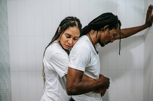 Side view sad loving African American female in white shirt standing behind and embracing depressed unhappy husband in bathroom