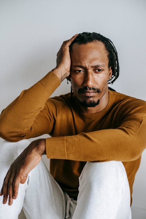Free Uncertain disappointed African American male in casual clothes touching head and looking away in frustration while sitting on floor against white wall Stock Photo
