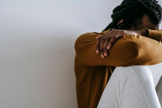 Crop anonymous depressed African American male embracing knees and covering face with arms while sitting on floor against white wall