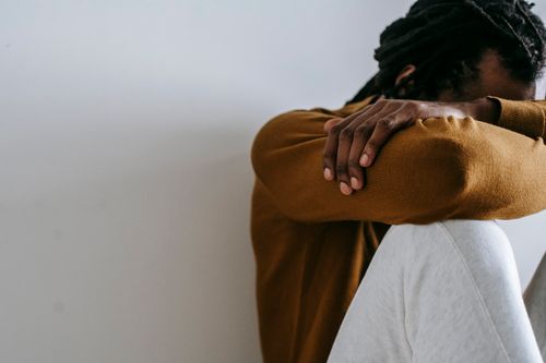 Free Crop anonymous depressed African American male embracing knees and covering face with arms while sitting on floor against white wall Stock Photo