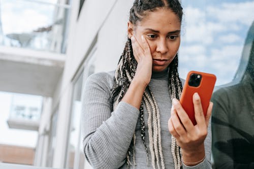 Free Crop serious African American female with dreadlocks touching cheek in contemplation and browsing mobile phone on street Stock Photo