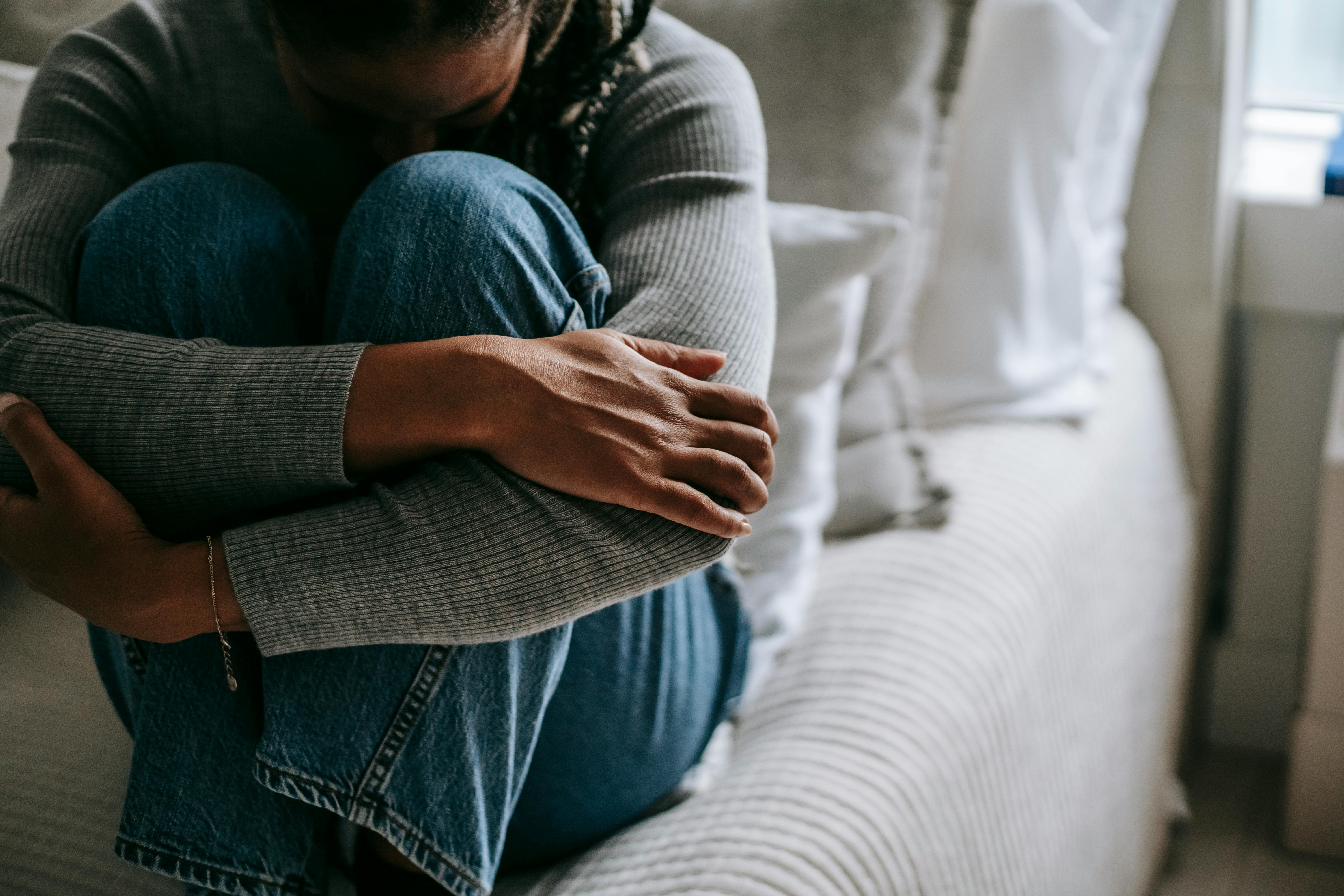 crop pitiful black woman embracing knees on bed