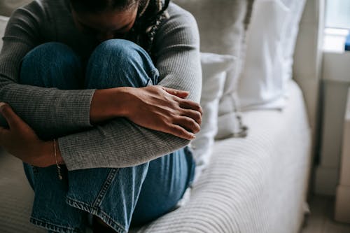 Crop pitiful black woman embracing knees on bed