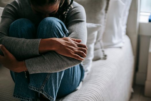 Crop pitiful black woman embracing knees on bed