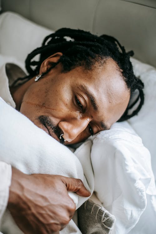 From above pondering stressed African American male hugging pillow and looking away in sad thoughts while lying on bed