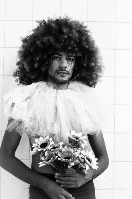 Black and white of man in costume of clown with Afro hairstyle holding bunch of flowers and looking at camera