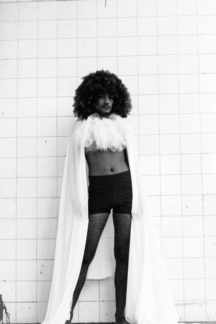 Black and white of freaky male in underwear and cloak standing near white tile wall and looking at camera