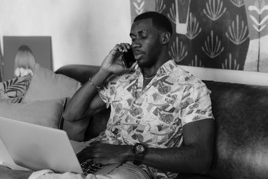 Black and white portrait of a man working from home, talking on the phone while using a laptop on the couch.