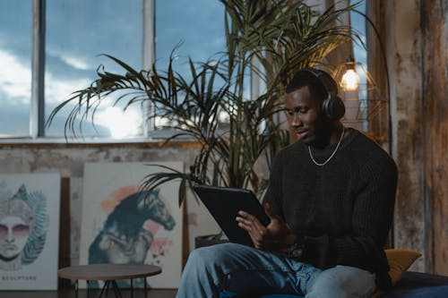 Man in Black Sweater and Blue Denim Jeans Using Digital Tablet and Headset 